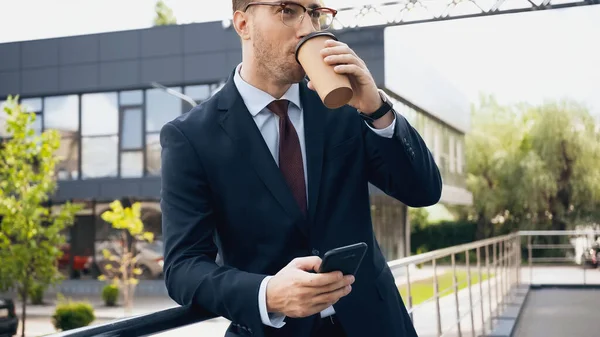 Homme d'affaires en lunettes et costume tenant smartphone tout en buvant du café à emporter — Photo de stock