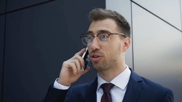 Businessman in glasses and suit talking on mobile phone near building — Stock Photo