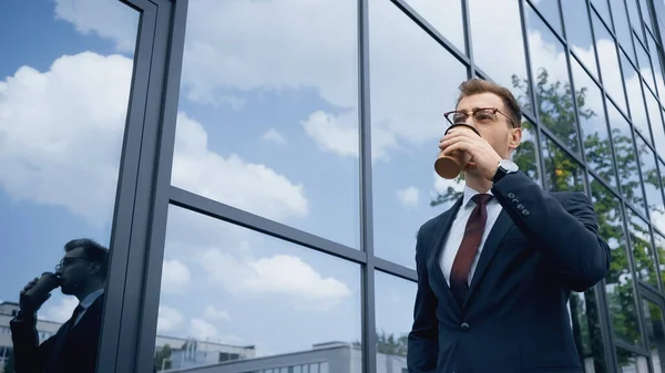 Бизнесмен в очках пьет кофе, чтобы подойти к зданию со стеклянным фасадом — стоковое фото