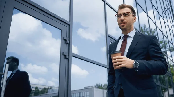 Businessman in glasses holding coffee to go near building with glass facade — Stock Photo