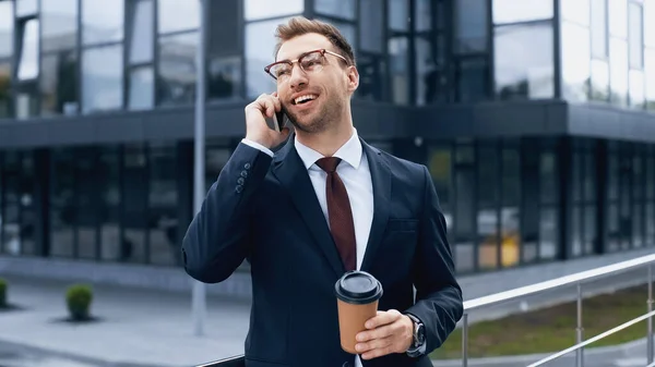 Homme d'affaires positif parlant sur téléphone portable et tenant du café pour aller à l'extérieur — Photo de stock