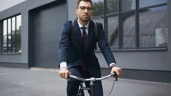 Homem de negócios de terno olhando para a câmera ao andar de bicicleta perto do edifício — Fotografia de Stock