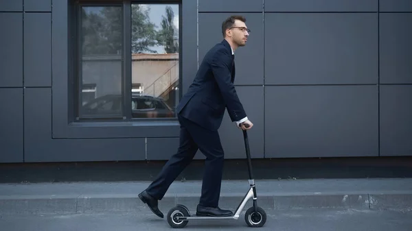 Full length of businessman in suit and glasses riding electric scooter near building — Stock Photo