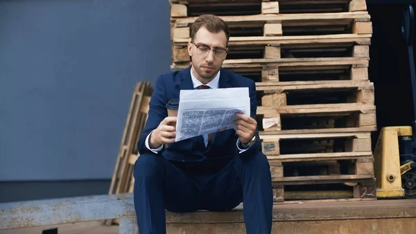 Homme d'affaires assis tout en tenant un café pour aller lire le journal à l'extérieur — Photo de stock