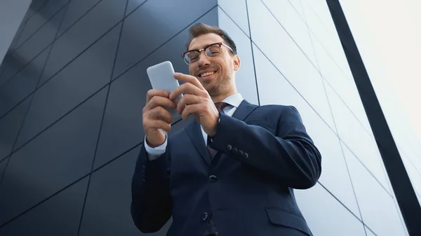 Vista de ángulo bajo de empresario alegre en gafas de mensajes de texto en el teléfono móvil fuera - foto de stock