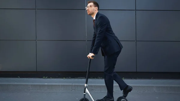 Full length of businessman in formal wear and glasses riding e-scooter near building — Stock Photo