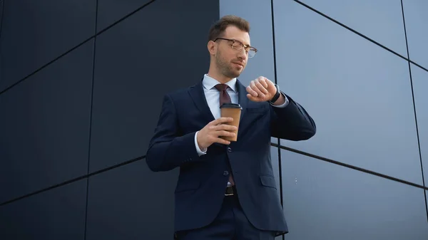 Бизнесмен в формальной одежде держит кофе, чтобы пойти и смотреть на наручные часы, пока ждет снаружи — стоковое фото