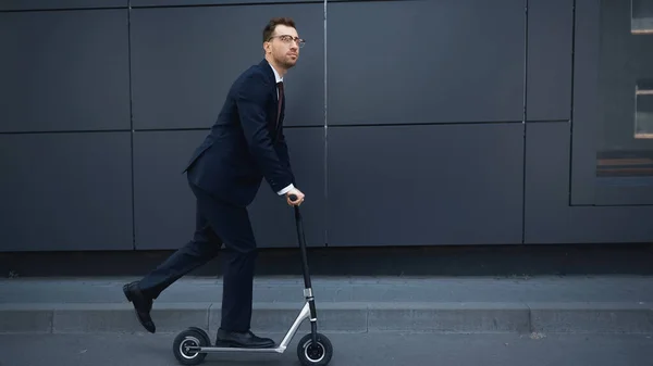 Pleine longueur de l'homme d'affaires en costume et lunettes équitation e-scooter près du bâtiment — Photo de stock