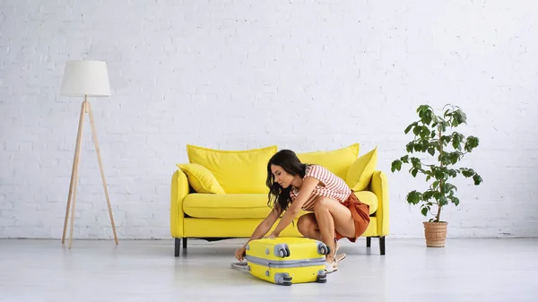 Mulher morena fechando saco de viagem amarelo perto do sofá na sala de estar — Fotografia de Stock