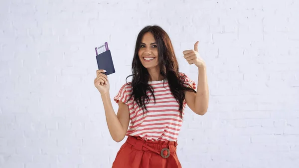 Mujer alegre sosteniendo pasaporte con billete de avión y mostrando el pulgar hacia arriba - foto de stock