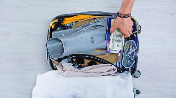 Vista recortada del hombre poniendo dinero en la bolsa de viaje con ropa y máscara de natación - foto de stock