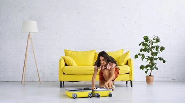 Брюнетка женщина сидит рядом желтый чемодан и диван в гостиной — стоковое фото
