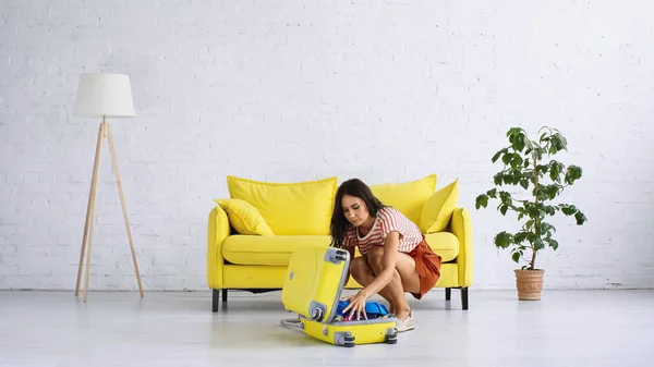 Brunette woman sitting and packing yellow suitcase near sofa in living room — Stock Photo