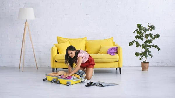 Morena mujer embalaje amarillo maleta cerca de sofá en sala de estar - foto de stock