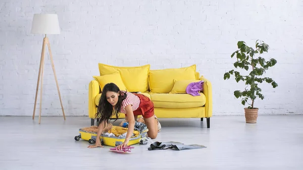Morena mujer llegar chanclas mientras embalaje amarillo maleta cerca de sofá en sala de estar - foto de stock