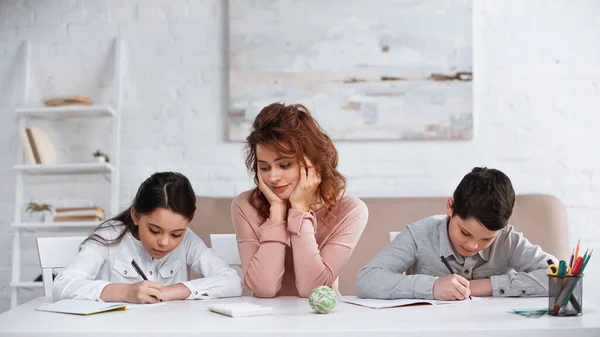 Mãe apoiando crianças escrevendo em cadernos durante os trabalhos de casa — Fotografia de Stock