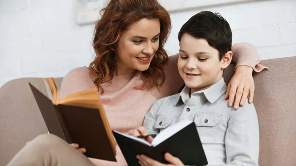 Sorridente madre che tiene libro vicino al figlio con notebook durante l'istruzione a casa — Foto stock