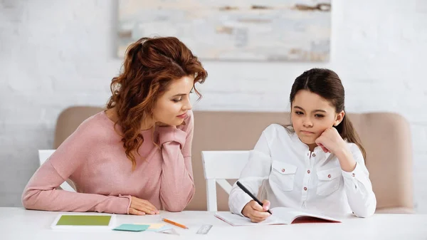 Pensive kid holding pen near notebook and mother at home — Stock Photo