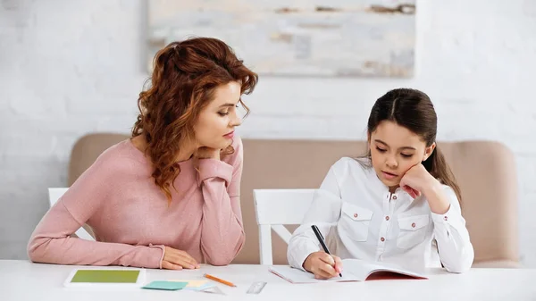 Mujer sentada cerca de un niño escribiendo en un cuaderno cerca de una tableta digital y notas adhesivas - foto de stock