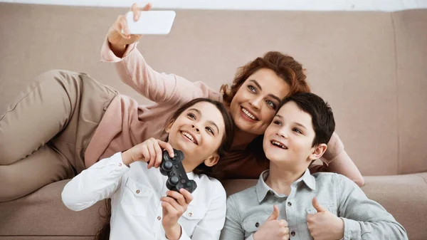 KYIV, UCRANIA - 15 DE ABRIL DE 2019: Mujer sonriente tomando selfie con una chica sosteniendo gamepad e hijo mostrando como gesto - foto de stock