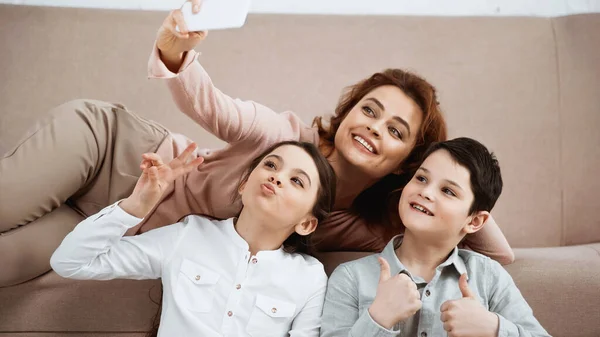 Mujer sonriente tomando selfie cerca de niños mostrando paz y gestos similares - foto de stock