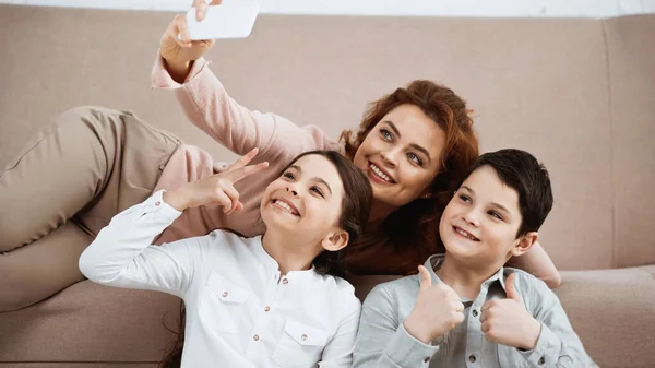 Crianças alegres gesticulando enquanto tomam selfie com mãe sorridente em casa — Fotografia de Stock