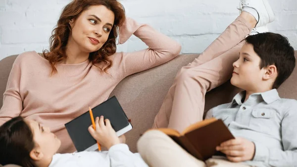 Mujer mirando a los niños con cuaderno y libro en primer plano borroso en el sofá - foto de stock