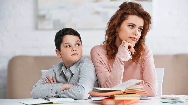 Hijo con los brazos cruzados mirando a la madre triste cerca de los libros en la mesa - foto de stock