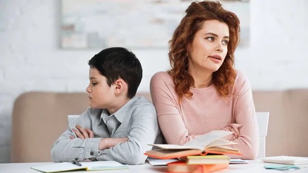 Mujer e hijo trastornados con los brazos cruzados sentados cerca de los libros en la mesa - foto de stock