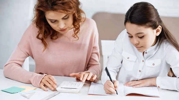 Mädchen schreibt auf Notizbuch neben Mutter und Taschenrechner zu Hause — Stockfoto