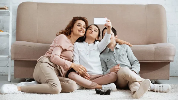 KYIV, UKRAINE -  APRIL 15, 2019: Smiling woman taking selfie with kids near gamepad on floor at home — Stock Photo