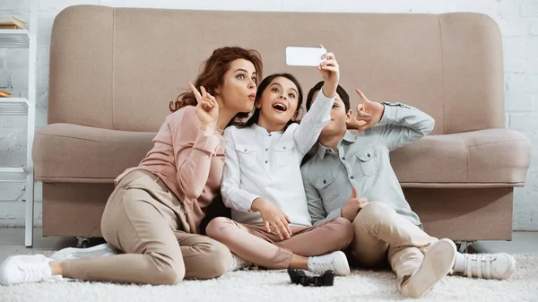 KYIV, UCRANIA - 15 DE ABRIL DE 2019: Familia tomando selfie en el teléfono inteligente cerca de joystick en el suelo en casa - foto de stock