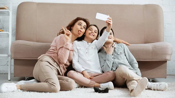KYIV, UCRANIA - 15 DE ABRIL DE 2019: Mujer positiva y niños tomando selfie cerca de joystick en casa - foto de stock