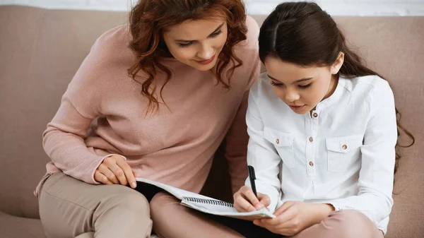 Écriture d'enfant sur un cahier près de la mère aidant pendant les devoirs — Photo de stock