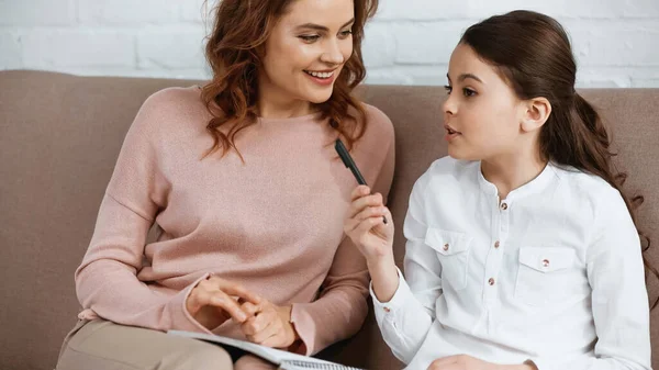 Girl holding pen and notebook near smiling mother on couch — Stock Photo