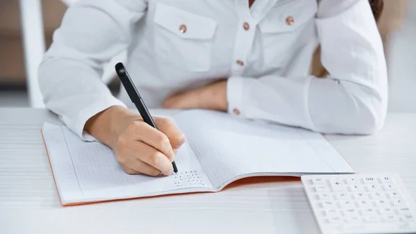 Cropped view of girl writing on notebook near calculator on blurred foreground — Stock Photo
