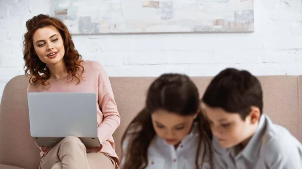 Lächelnde Frau mit Laptop, die zu Hause Kinder im verschwommenen Vordergrund betrachtet — Stockfoto