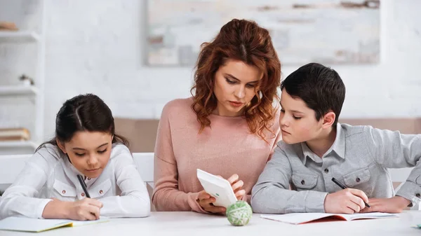 Mulher mostrando calculadora para filho perto filha escrevendo no notebook em casa — Fotografia de Stock