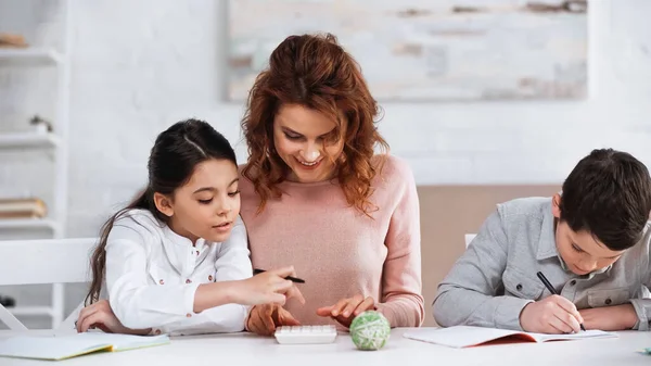 Smiling woman sitting near daughter using calculator and son writing on notebook — Stock Photo