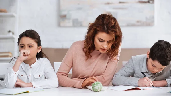 Mulher usando calculadora perto da filha pensativa e filho escrevendo no notebook em casa — Fotografia de Stock