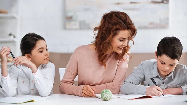 Mujer sonriente sentada cerca de su hijo escribiendo en el cuaderno y su hija en casa - foto de stock