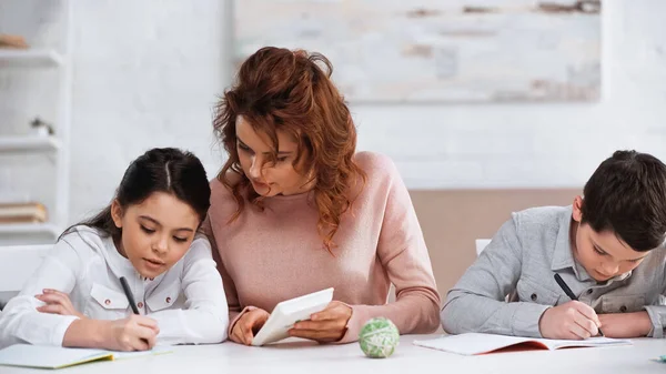 Mulher segurando calculadora perto de crianças escrevendo em cadernos em casa — Fotografia de Stock