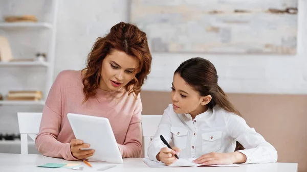 Woman showing digital tablet near daughter writing on notebook — Stock Photo