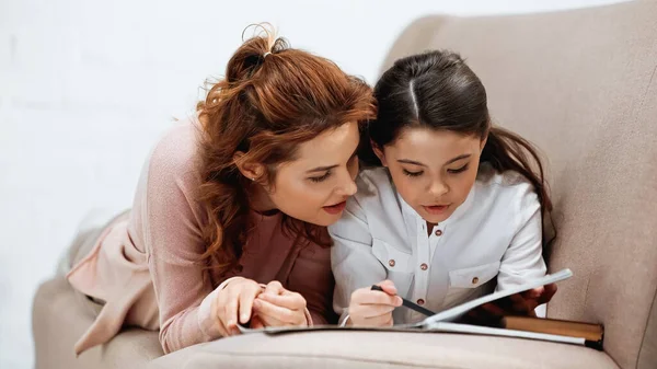 Donna sdraiata vicino a figlia che fa i compiti e laptop sul divano — Foto stock