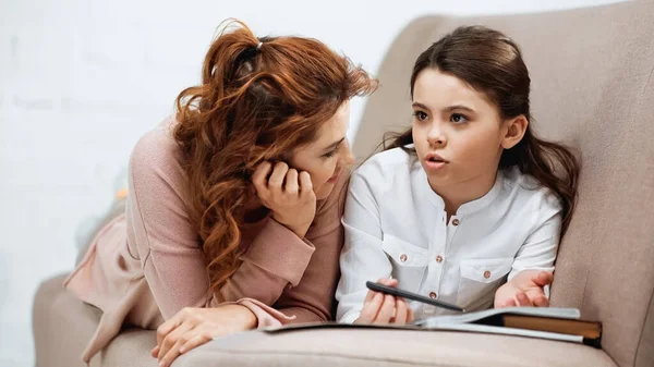 Chica hablando cerca de la madre y el ordenador portátil mientras hace la tarea en el sofá - foto de stock