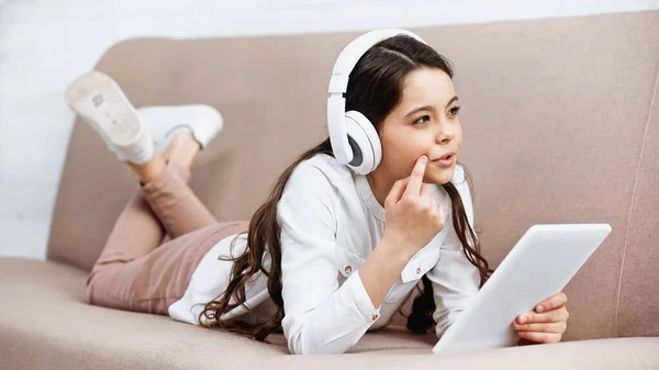 Chica preadolescente pensativa en auriculares que sostienen la tableta digital en el sofá en casa - foto de stock