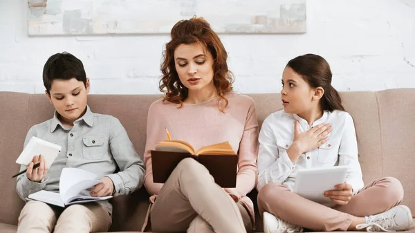 Mujer leyendo libro cerca de los niños con tableta digital y portátil - foto de stock