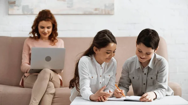 Bambini sorridenti che scrivono su notebook vicino alla madre usando laptop su sfondo sfocato in soggiorno — Foto stock