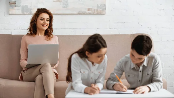 Smiling teleworker looking at kids doing homework on blurred foreground — Stock Photo