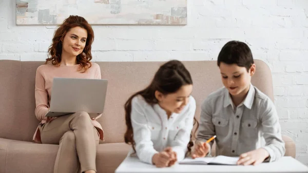 Woman with laptop looking at kids with notebook doing homework on blurred foreground — Stock Photo